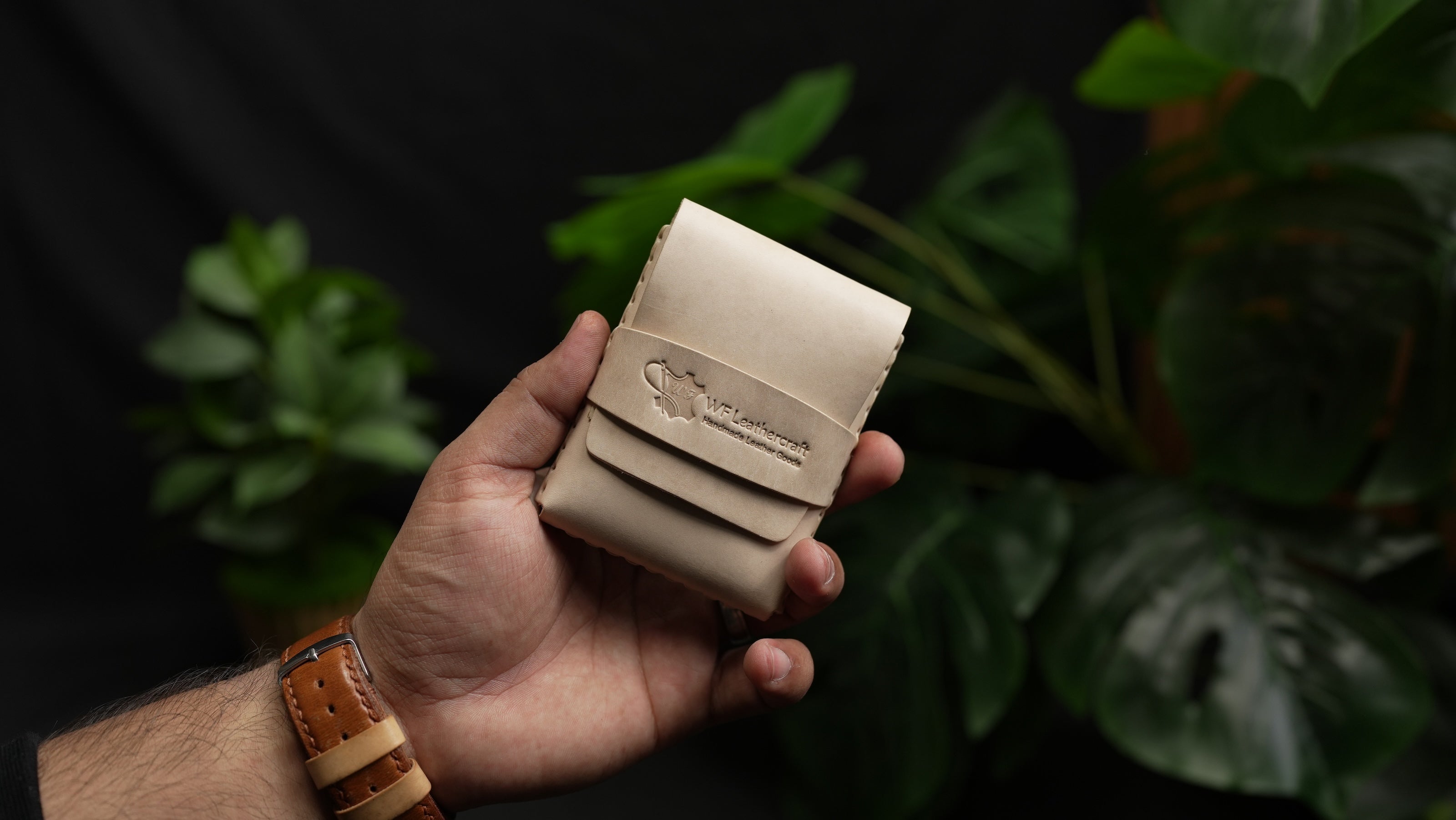Hand holding a beige leather wallet with a brand logo against a dark background with green plants.