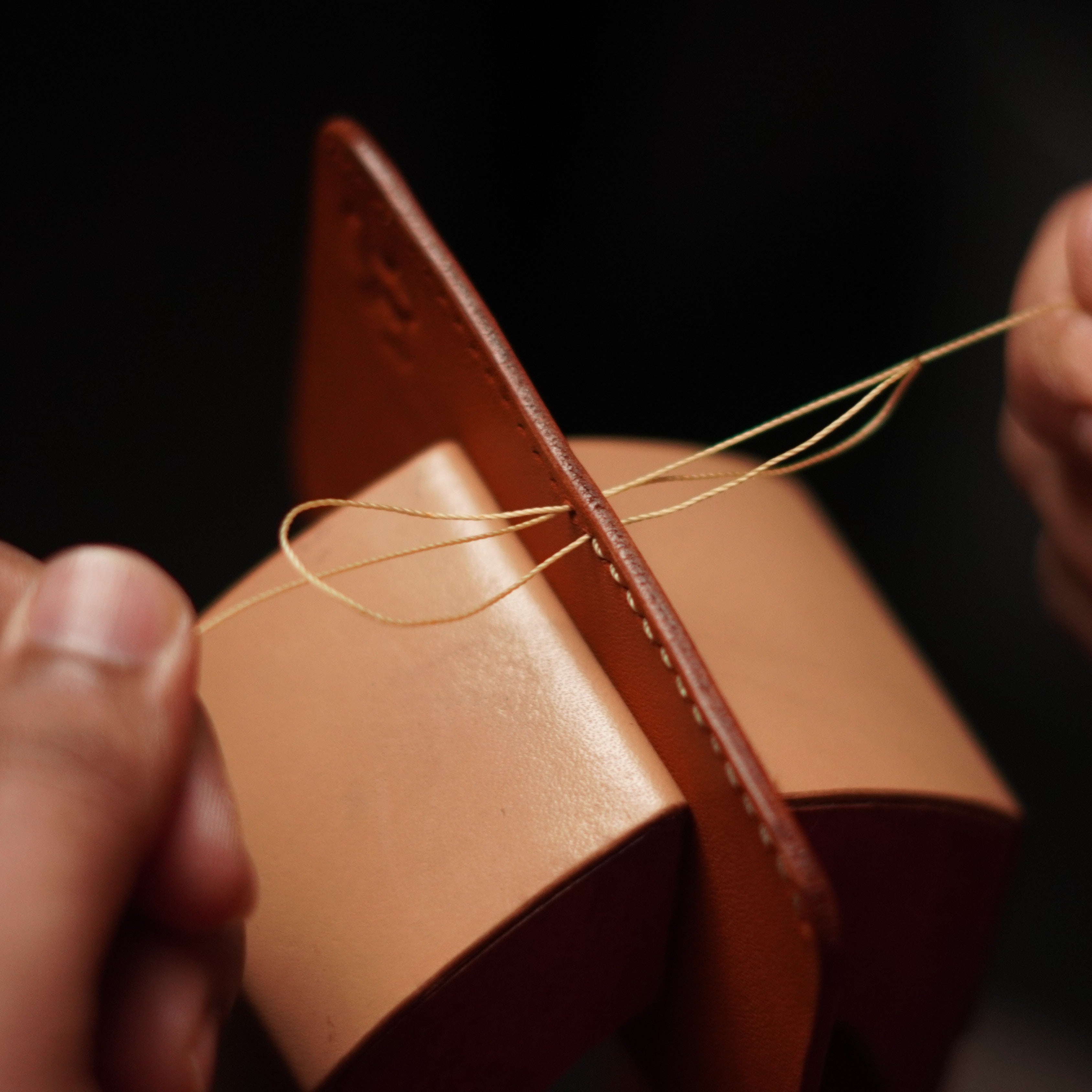 Brown leather wallet with a thread being threaded through it against a dark background
